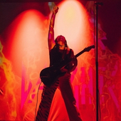 Charlene Kaye on stage in dark clothing, red lights behind her. She is holding her guitar and lifting her arm up in the air.